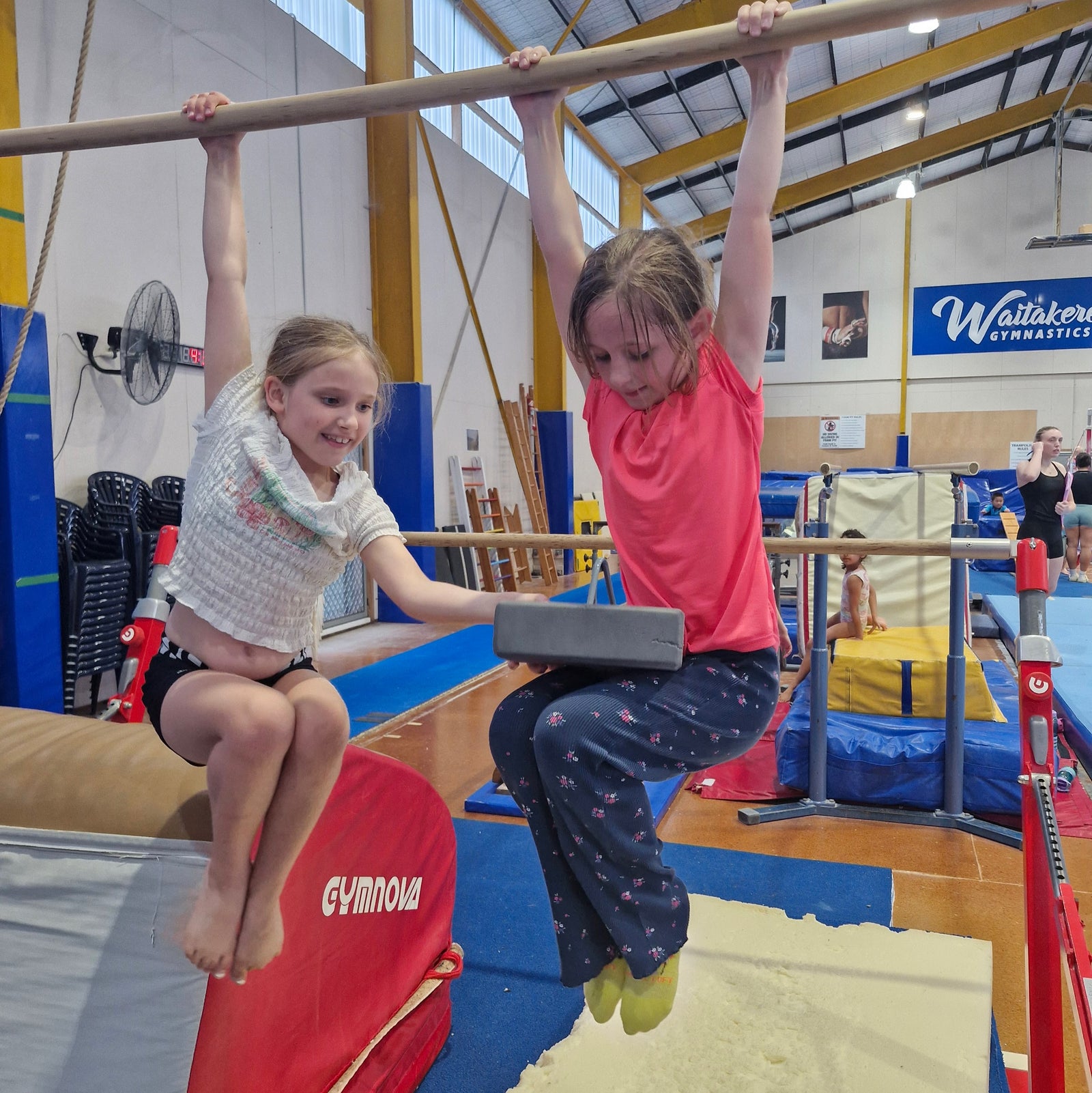 Two girls performing a tuck hold on high bar during gymnastics class