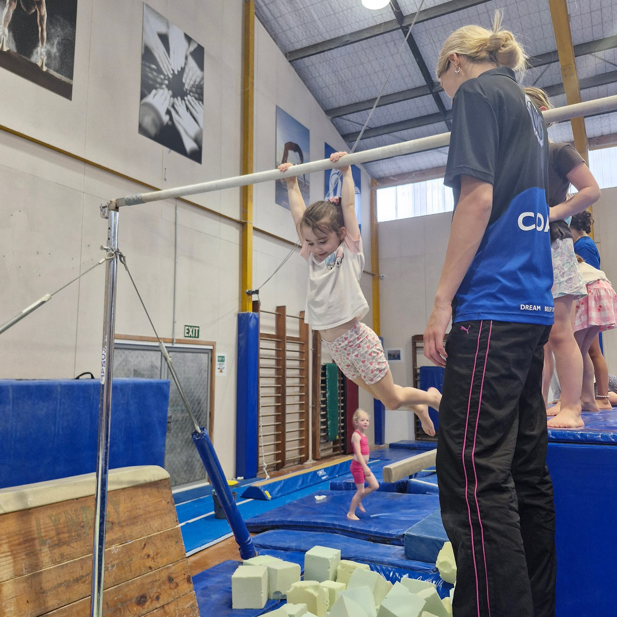 Girl wearing shorts and T-shirt swinging on high bar over foam pit during gymnastics class